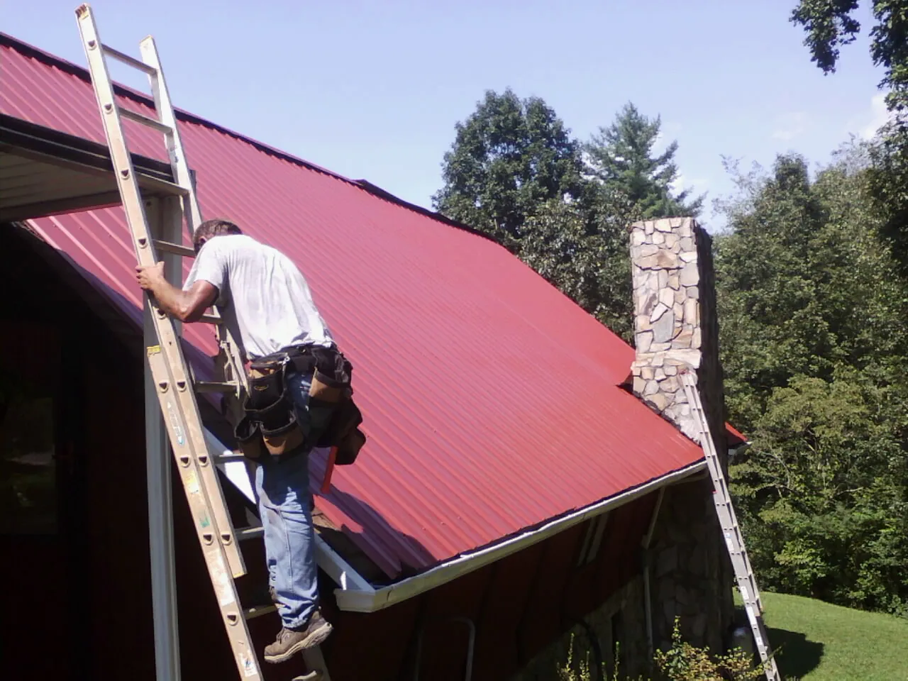 Roofer on a ladder beside a red metal roof and stone chimney — Soffit & Fascia Repair by Roofing Brothers in Schuylkill Haven